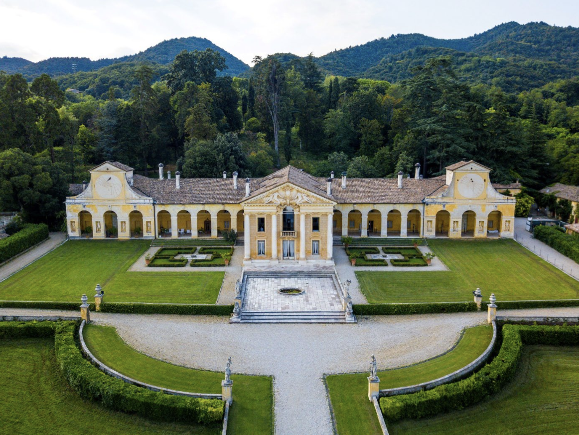 Luftaufnahme einer historischen Villa mit symmetrischem Garten, Wegen und bewaldeten Hügeln im Hintergrund.